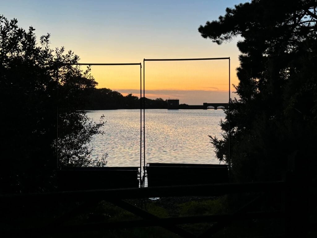 Sunset over a lake with silhouetted trees and fence.