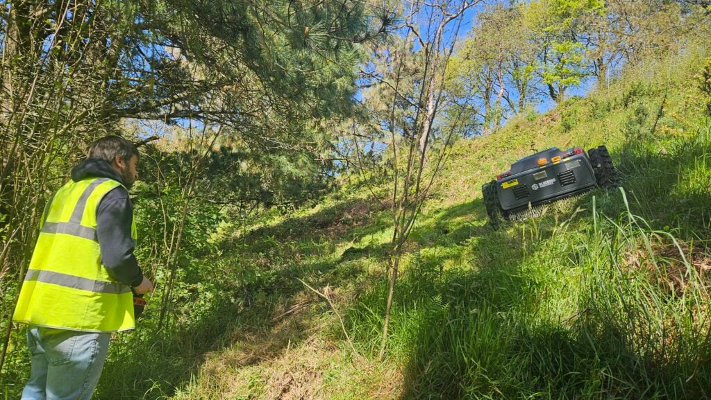 Man controls robot mower on grassy hill.