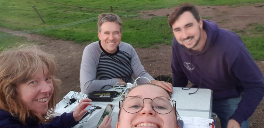 Group smiling outdoors with electronics on table.